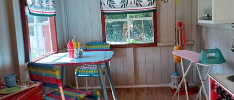 A cozy room with wooden walls, a wooden ceiling, a window with a view of greenery, a table with chairs, a red cabinet, and a shelf with items.