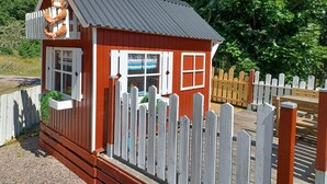 Un petit bâtiment en bois rouge, avec une clôture blanche en piquets, un toit en métal et une fenêtre dotée de volets blancs.