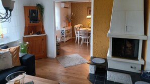 Un salon chaleureux avec une cheminée, un coin repas avec une table et des chaises, et une cuisine équipée de placards et agrémentée d’une plante.
