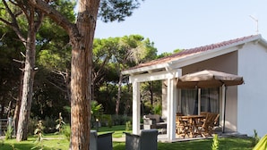 A covered patio area with outdoor furniture, a white building with a tiled roof, and surrounding greenery.