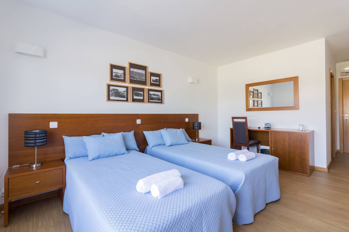 Habitación de hotel con dos camas, un cabecero de madera, un escritorio, un espejo y cuadros enmarcados en la pared.