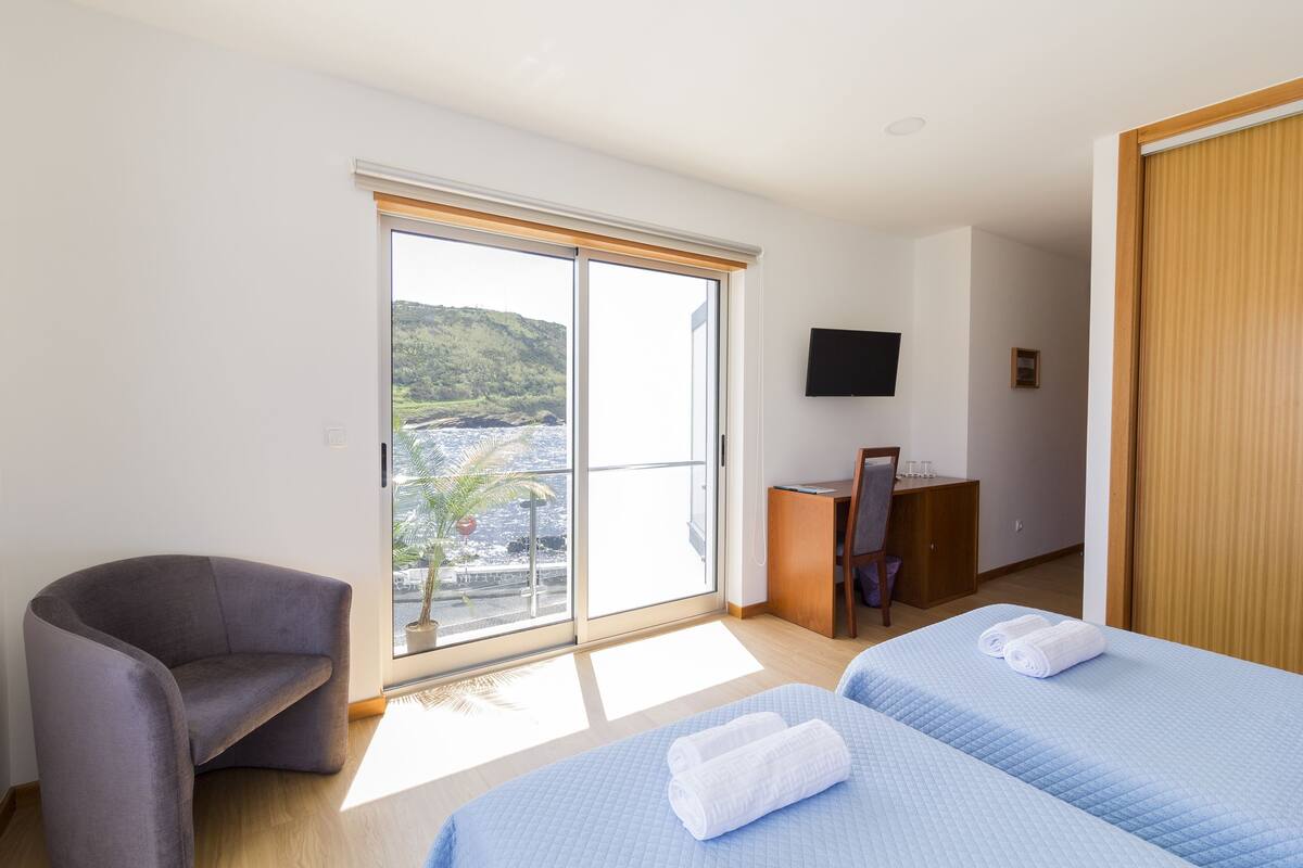 Habitación de hotel con cama, escritorio, silla, televisor y ventana grande con vistas a una ladera.