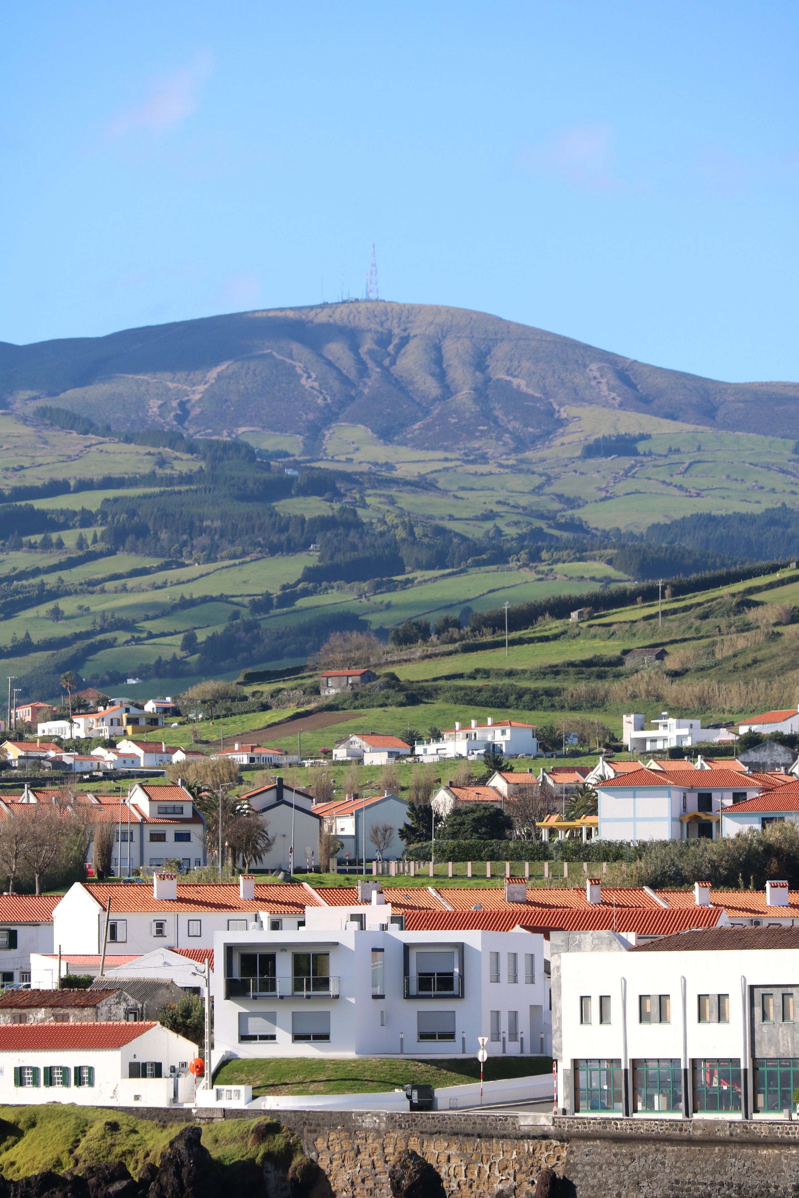 A residential area with modern houses, a stone wall, and a hill with a tower in the background.