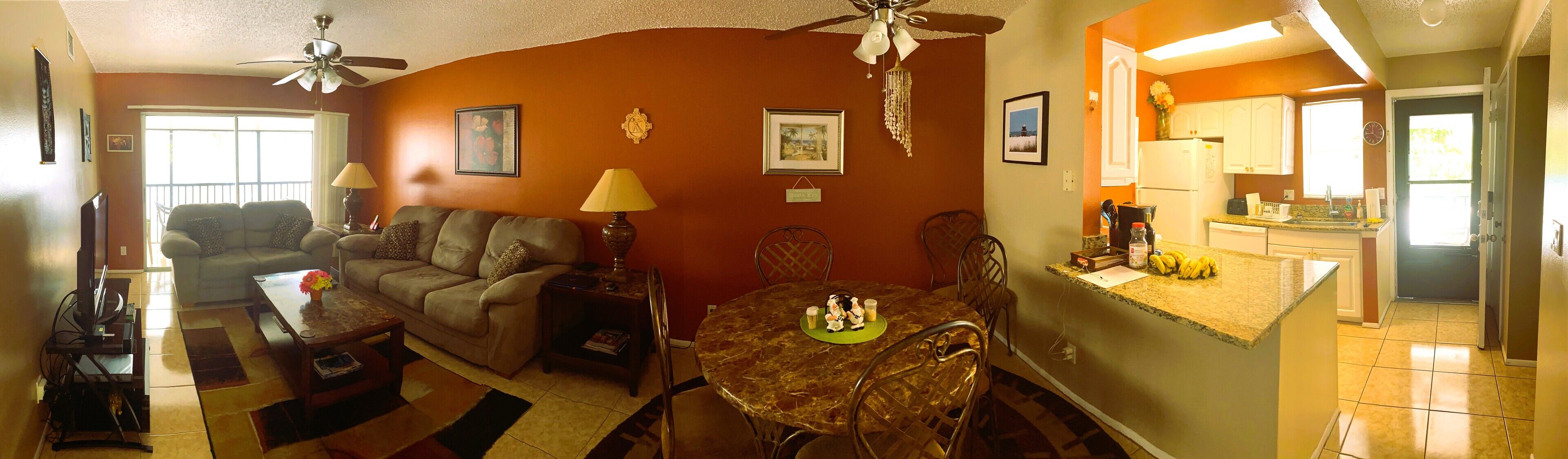 A living room with a sofa, coffee table, and dining table with chairs. There is a kitchen area with a refrigerator and a window.