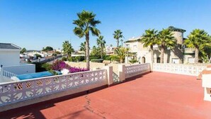 A rooftop terrace with a red floor, palm trees, and a white building with a balcony.