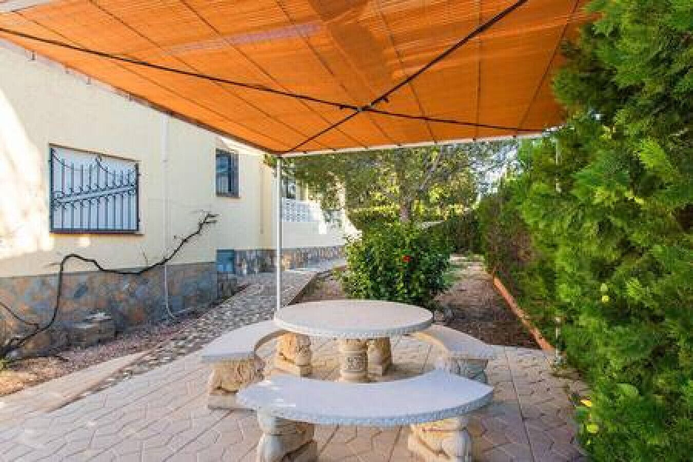An outdoor patio with a round table and chairs, a covered area with a brown canopy, and a stone pathway.