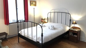 A bedroom with a metal-framed bed, a bedside table with a lamp, a radiator, and a window with red curtains. - ST-JORIOZ, maison de famille, 500m du Lac, 10M de la piste cyclable, 10pers (Saint-Jorioz)