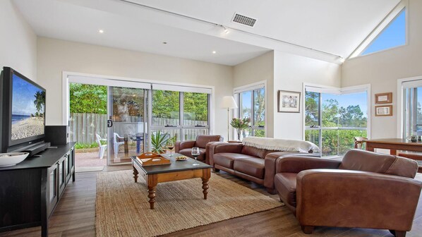 A spacious living room with a television, leather sofas, a coffee table, and large sliding glass doors leading to a balcony with a view of greenery.