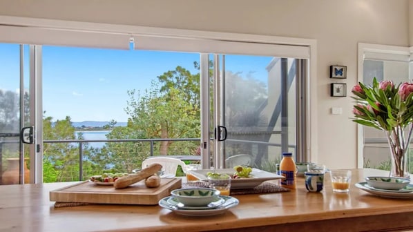 Un comedor con vista a un cuerpo de agua y vegetación, una mesa de madera con comida y bebidas, y un jarrón con flores.