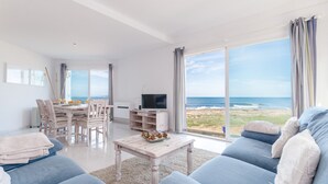 A spacious living room with a large window overlooking the beach, a dining area with a table and chairs, and a flat-screen TV on a wooden stand. - YourHouse Solmarina Son Serra (Son Serra de Marina)
