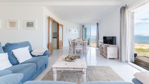 A modern living room with a blue sofa, a wooden coffee table, and a TV. There is a dining area with a table and chairs, and a view of the sea through large windows. - YourHouse Solmarina Son Serra (Son Serra de Marina)