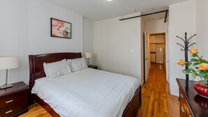 Una cama bien hecha con una colcha blanca y almohadas, una mesita de noche con una lámpara, una obra de arte en la pared y un pasillo que conduce a una zona de cocina.