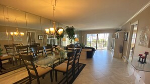 A spacious living area with a glass dining table, comfortable seating, and a large window offering a view of the outdoors.