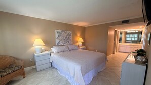 A bedroom with a large bed, two bedside lamps, a wicker chair, a white dresser, and a bathroom visible through an open door.