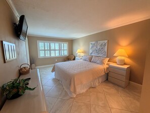 Quarto com cama, luminárias de cabeceira, televisão, janela e obra de arte na parede.