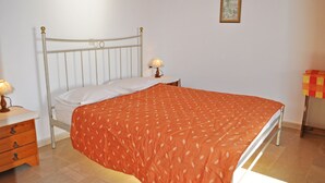 Een slaapkamer met een bed met metalen frame, wit beddengoed, een oranje bedsprei met patroon, een nachtkastje met een lamp, en een klein nachtkastje met een lamp.