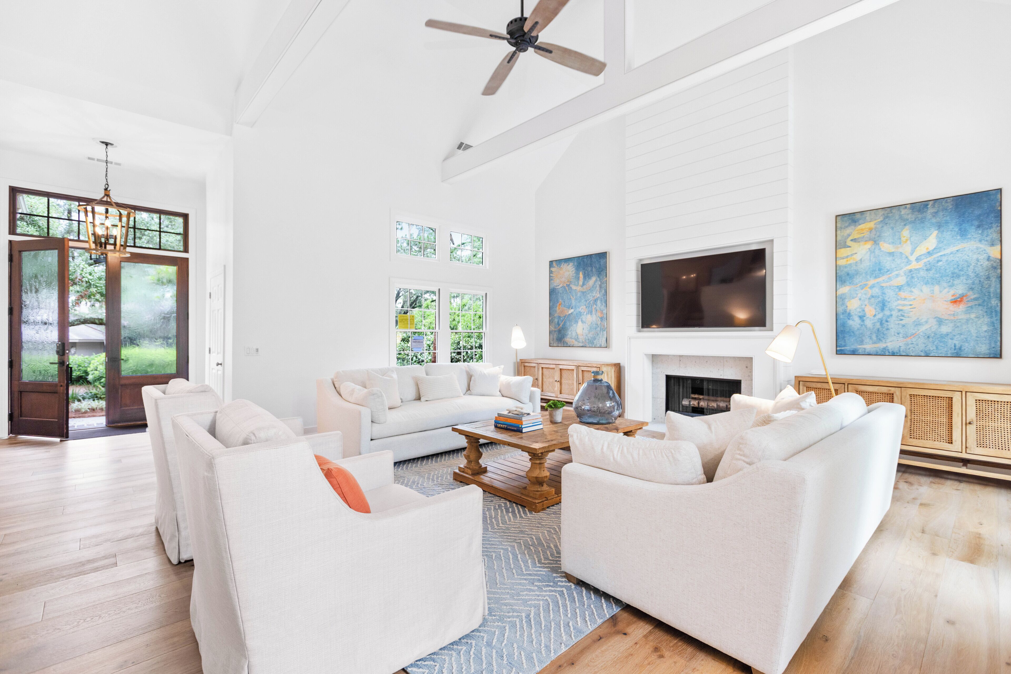 A spacious living room with a white sofa set, a coffee table, and a fireplace. There are large windows and a ceiling fan.