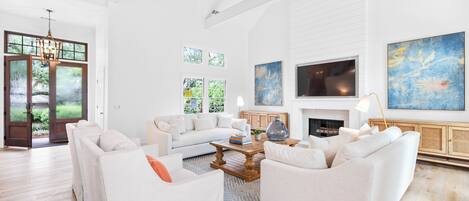 A spacious living room with a white sofa set, a coffee table, and a fireplace. There are large windows and a ceiling fan.