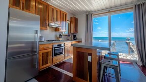 A modern kitchen with wooden cabinets, stainless steel appliances, and a view of the ocean through large windows. - Box House Boca Ciega - Unique Beachfront Container Home (San Pedro)