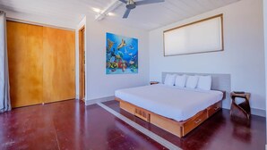 A bedroom with a bed, a ceiling fan, a painting, and a wooden door. - Box House Boca Ciega - Unique Beachfront Container Home (San Pedro)