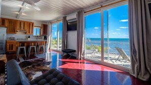 A spacious living area with a large window overlooking the ocean, a kitchen with wooden cabinets, and a ceiling fan. - Box House Boca Ciega - Unique Beachfront Container Home (San Pedro)