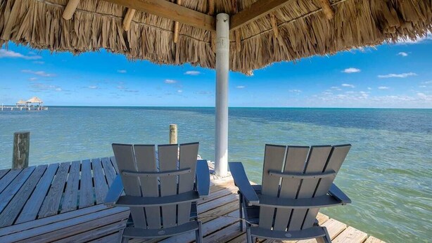 A wooden deck with two chairs facing a thatched roof structure overlooking the ocean. - Box House Boca Ciega - Unique Beachfront Container Home (San Pedro)