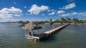 Ein Steg, der sich in ruhiges Wasser erstreckt, mit einem Gebäude mit Strohdach am Ende, einem angedockten Boot und einem Haus im Hintergrund.