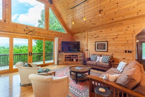 A spacious living room with a large TV, comfortable seating, and a balcony with a scenic view.