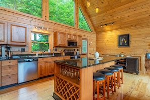 A modern kitchen with wooden cabinetry, a central island with bar stools, and large windows offering a view of the forest.
