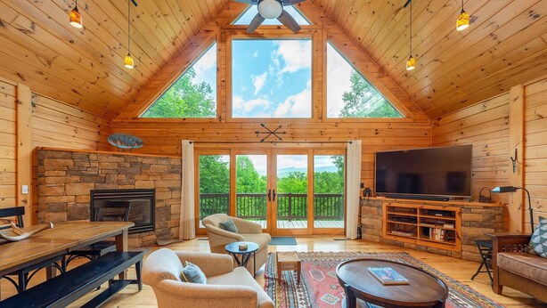 Interior de una cabaña de troncos con chimenea de piedra, muebles de madera, televisor grande y balcón con vistas a la vegetación.