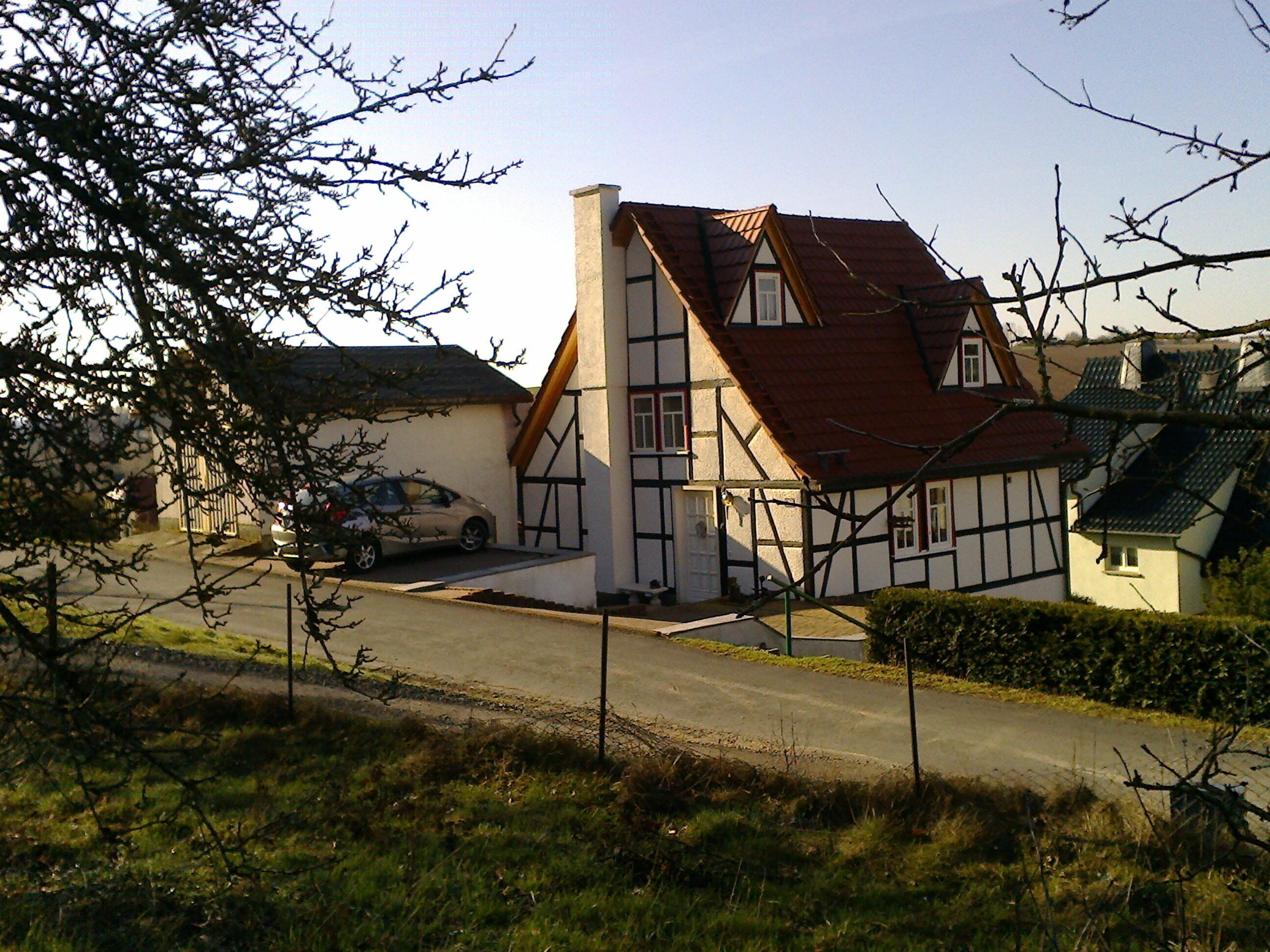 A traditional half-timbered house with a steeply pitched roof, a car parked in front, and a clear sky.