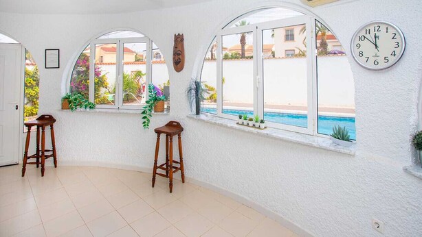 Ein Zimmer mit zwei hölzernen Hocker, einem Deckenventilator, einer Wanduhr und Blick auf einen Pool durch große Fenster.