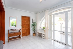 Un couloir lumineux avec une porte en bois, un banc, une plante en pot et une grande porte vitrée donnant sur l’extérieur.