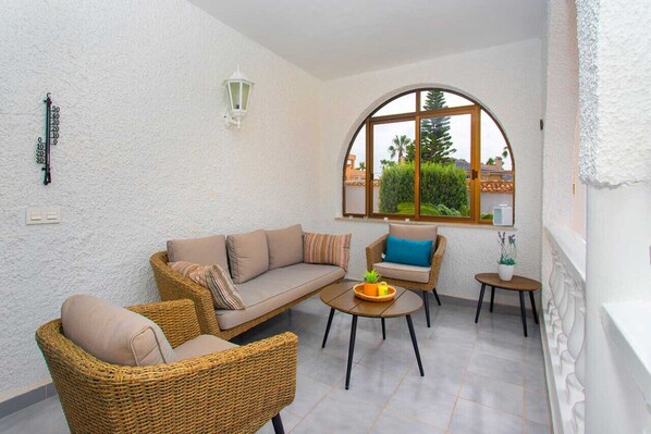 A sunlit patio with wicker furniture, a small table with a plant, and a view of greenery through a large arched window.