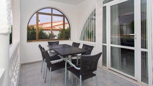 Eine überdachte Terrasse mit Tisch und Stühlen, ein großes Fenster mit Blick auf Gebäude und eine Glastür.
