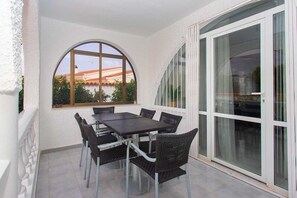 Une terrasse couverte avec une table et des chaises, une grande fenêtre donnant sur les bâtiments et une porte-fenêtre coulissante.