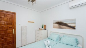 Ein Schlafzimmer mit Holztür, weißem Schminktisch, einem Bett mit gemusterten Textilien und einer Wand-Klimaanlage, sowie einem Bild mit Strandmotiv.