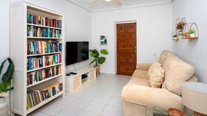 Ein Wohnzimmer mit einem beigefarbenen Sofa, einer weißen Bücherwand voller Bücher, einem Flachbildfernseher, einem Deckenventilator und einer Holztür.