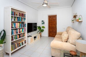 Un salon avec un canapé beige, une bibliothèque blanche remplie de livres, un téléviseur à écran plat, un ventilateur de plafond et une porte en bois.