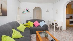 A living room with a grey sofa, colorful cushions, a glass coffee table, and a white dining area with a small table and chairs.