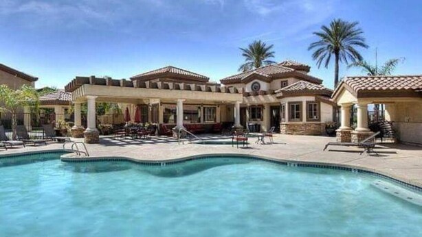A pool area with a clear blue pool, lounge chairs, and a building with a tiled roof and columns. - Resort Style Vacation Condo (Mesa)