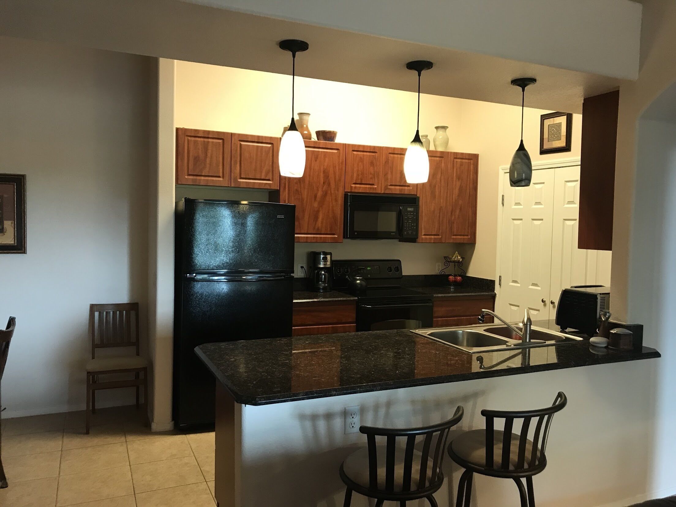 Une cuisine avec des meubles de rangement en bois, un réfrigérateur noir, un four à micro-ondes, une cuisinière et deux lampes suspendues.