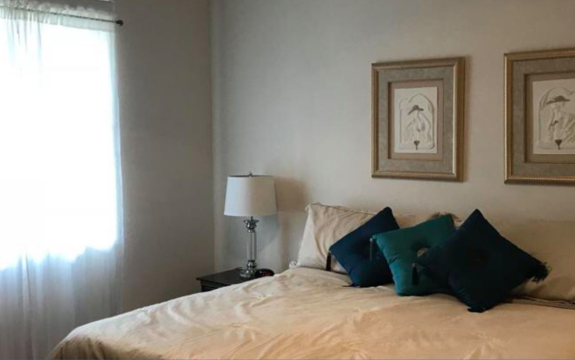 Un dormitorio con cama, almohadas, una mesita de noche, lámpara y dos cuadros en la pared.