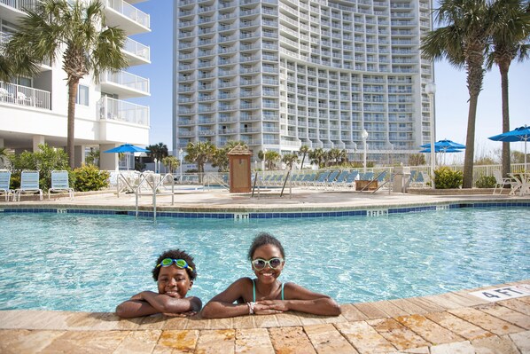 A swimming pool with two people in the water, surrounded by lounge chairs and palm trees, with a tall hotel building in the background.