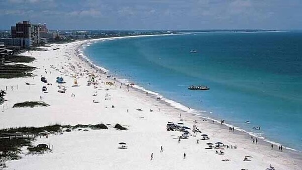 Uma praia com areal dourado, alguns edifícios ao fundo e um céu azul límpido.