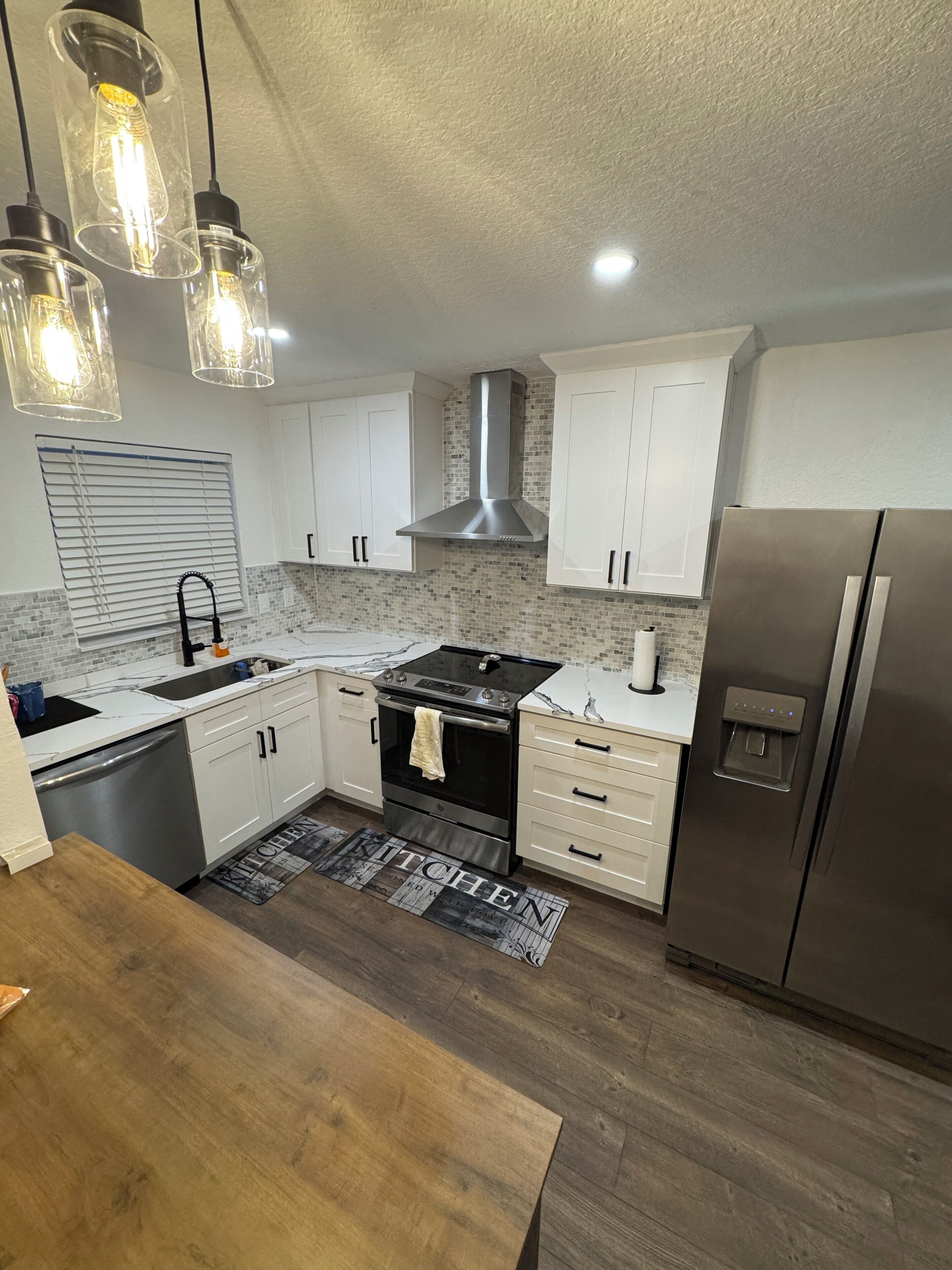 A modern kitchen with white cabinets, a stainless steel refrigerator, a built-in oven, and pendant lights.