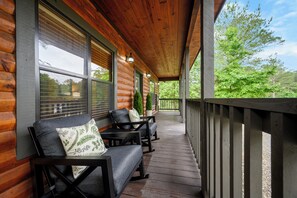 A covered porch with wooden railings, a seating area with two sofas and a table, and a view of greenery.