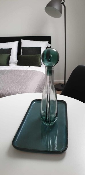 Ein Bett mit schwarzem Kopfteil und weißer Bettwäsche, einem grünen Kopfkissen und einer klaren Glasvase mit grünem Deckel auf einem weißen Tisch.