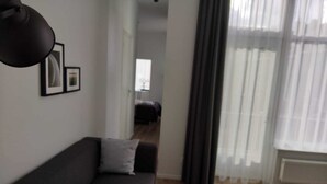 Una sala moderna con un sofá oscuro, una mesita auxiliar y una lámpara de pie negra. Un ventanal con cortinas translúcidas permite el paso de la luz natural.
