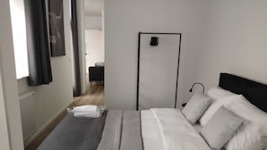 Una cama bien tendida con sábanas blancas y un edredón gris, un cabecero negro y una mesita de noche con una lámpara.